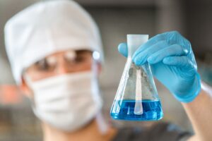 Laboratory technician in protective gloves and mask holding a flask of blue liquid during formulation testing, representing the quality-control process behind Germany's OEM sunscreen and sun-care manufacturing.